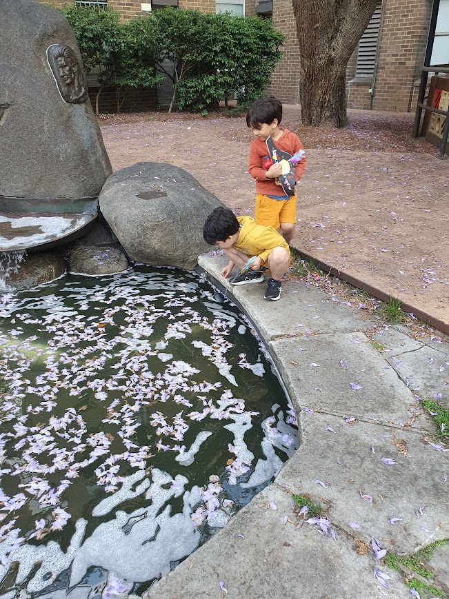 Two Pasifika boys at UNSW’s Morven Brown courtyard peer into a stone fountain/pond. One boy crouches at the edge while the other stands holding pirate hat. Purple jacaranda petals cover the water and path.